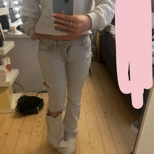 Ljusgrå bootcut jeans från mango  - Säljer ett par ljusgrå bootcut jeans från mango med slitna detaljer och hål på benen. Jeansen har låg midja och klassisk femficksdesign. Perfekta för dig som gillar en avslappnad och trendig look.