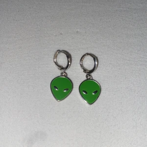 Örhängen med gröna alienhuvuden - Coola örhängen med gröna alienhuvuden som hänger i små silverfärgade ringar. Perfekt accessoar för dig som gillar unika och lekfulla smycken med en twist av sci-fi.