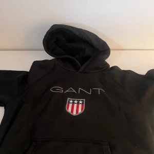 Svart hoodie från GANT med logga - Svart hoodie från GANT med broderad logga och sköld på bröstet. Klassisk huva och känguruficka framtill. Snygg och enkel design med vita och röda detaljer på loggan. Perfekt för dig som gillar stilrena streetwear-plagg.