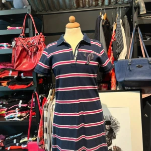 Randig marinblå kortklänning  - Snygg kortklänning från Polo Ralph Lauren i marinblått med vita och rosa ränder. Klassisk pikékrage, korta ärmar och broderad P-logga på bröstet. Klänningen har knappar framtill och är gjord i mjuk bomull. Perfekt för en chill och sportig look.