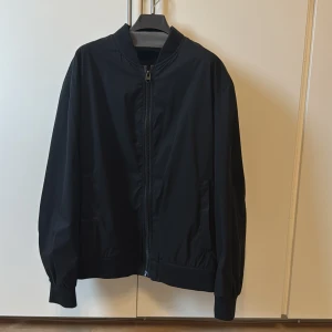 Svart bomberjacka från Zara - Säljer en svart bomberjacka från Zara i storlek XL. Jackan har klassisk bombermodell med dragkedja framtill, ribbade muddar och krage. Tillverkad i ett lätt och slätt syntetmaterial som känns skönt att bära. Perfekt för dig som gillar stilren och enkel look.