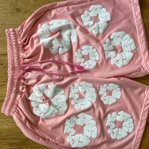 Rosa DenimTears shorts  - Unika rosa shorts med vita grafiska tryck över hela plagget. De har elastisk midja med rosa snörning och en bakficka. Modellen är loose fit och materialet känns mjukt och bekvämt, perfekt för sommardagar.