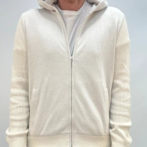 Beige stickad hoodie med dragkedja - Snygg beige hoodie med dragkedja framtill och huva. Tröjan är stickad med ribbade muddar och ärmar, samt har fickor på sidorna. Perfekt för en chill och stilren look.