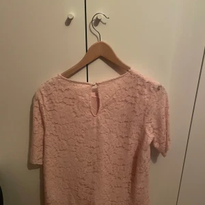 Ljusrosa spetsblus med kort ärm - Superfin ljusrosa blus från Kappahl i spets med korta ärmar och rund halsringning. Blusen har ett litet sprund med knapp i nacken och är helfodrad undertill. Perfekt för dig som gillar romantisk och feminin stil.