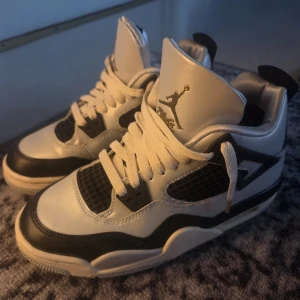 Nike Air Jordan 4 svart/vit sneakers - Säljer ett par Nike Air Jordan 4 sneakers i svart och silver med guldiga detaljer. Skorna har klassisk snörning, meshpaneler och den ikoniska Jumpman-loggan på häl och plös. Tillverkade i skinn och syntet, med en robust platt sula och rund tå. Perfekta för dig som vill sticka ut. Fick dom på julafton men dom var för små för mig, äkta såklart. Köpta från zalando för 2000kr
