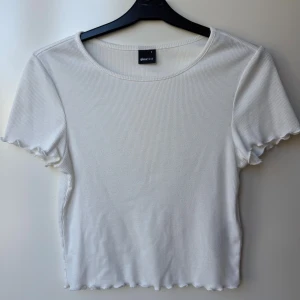 Vit ribbad topp från Gina Tricot - Superfin vit ribbad topp från Gina Tricot med korta ärmar och vågig kant nertill och vid ärmslut. Modellen är croppad och har en rund halsringning. Perfekt att matcha med jeans eller kjol för en clean look.