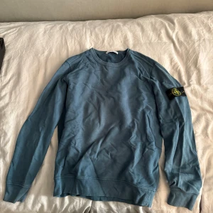 Blå sweatshirt från Stone Island - Säljer en blå långärmad sweatshirt från Stone Island med den klassiska loggan på vänster ärm. Tröjan har rund halsringning och är gjord i mjuk bomull, perfekt för en chill och stilren look.