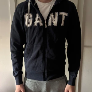 Marinblå hoodie från GANT med dragkedja - Marinblå hoodie från GANT med stora vita bokstäver framtill. Tröjan har huva med snörning, hel dragkedja och två fickor. Tillverkad i mjuk bomull och har ribbade muddar vid ärmslut och nederkant. Perfekt för chill dagar.