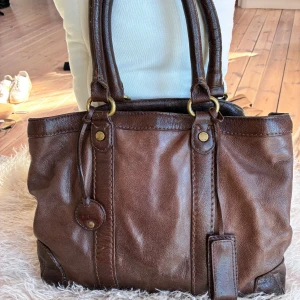 Brun handväska i skinn från Tommy Hilfiger - Säljer en snygg brun handväska i äkta skinn från Tommy Hilfiger. Väskan har dubbla handtag, mässingsfärgade metalldetaljer och flera fack inuti med dragkedja. Perfekt storlek för att få plats med allt du behöver till skolan eller stan. Liten fickspegel inklädd i skinn hängande på framsidan.