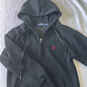 Svart hoodie från Polo Ralph Lauren - Svart hoodie från Polo Ralph Lauren med dragkedja och huva. Klassisk röd broderad logga på bröstet och snörning i huvan. Tillverkad i mjuk bomull, perfekt för chill dagar eller när du vill ha en clean look.