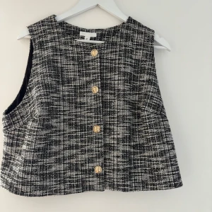 Rutig väst från H&M med guldknappar - Snygg rutig väst från H&M i svart och vitt med exklusiva guldknappar framtill. Västen har en boxig passform och är tillverkad i ett strukturerat tyg som ger en lyxig känsla. Perfekt att styla över en skjorta eller t-shirt för en trendig look. Den är som ny! 