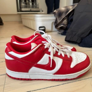 Nike Dunk Low Saint John University Red - Nike Dunk Low sneakers i rött och vitt skinn med klassisk Swoosh-logga på sidorna. Lågt skaft, vit snörning och perforerad tåbox för extra ventilation. Röd yttersula och innerfoder. Perfekt för dig som gillar streetwear och vill sticka ut. Gott skick, köpt i Gbg, kan skickas eller hämtas :)