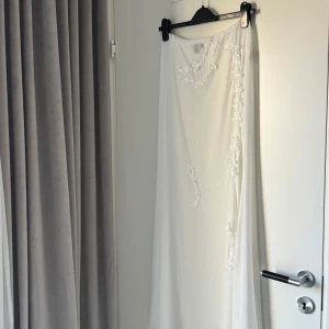 Vit strand kjol med blommig spets - Säljer en superfin vit oanvänd långkjol med med slits, mesh tyg och broderade blommor i spets längs kanten och framtill. Kjolen har en flowy och luftig känsla, perfekt för stranden passar även för strlk S finns i Vasastan 