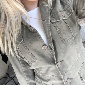 Grön overshirt från SIS by Sisters - Snygg grön overshirt från SIS by Sisters med fyra stora fickor framtill och knappar hela vägen. Jackan har lång ärm med knappdetaljer vid mudden och är tillverkad i mjuk bomull. Perfekt för dig som gillar utility-vibe och vill ha en chill look.