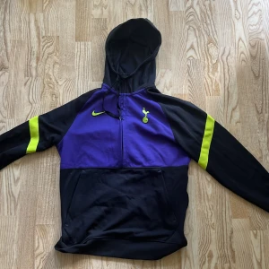 Nike Tottenham hoodie lila/svart S - Nike Tottenham hoodie i lila och svart med neongula detaljer på ärmarna. Tröjan har huva med snörning, stor ficka framtill och halv dragkedja. Dri-FIT-material som andas, perfekt för fotboll eller chill. Snygg Tottenham-logga på bröstet.