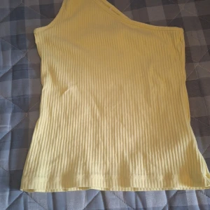 Gul ribbad oneshoulder topp Name It - Snygg ljusgul ribbad topp från Name It i oneshoulder-modell. Toppen är ärmlös och har en asymmetrisk skärning med bred axelrem på ena sidan. Tillverkad i mjuk bomull, perfekt för sommarens alla tillfällen.