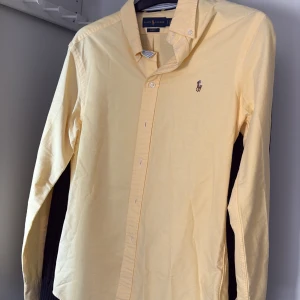 Gul skjorta från Ralph Lauren - Säljer en stilren gul skjorta från Ralph Lauren i slim fit-modell. Skjortan har klassisk krage, knappar framtill och den ikoniska broderade loggan på bröstet. Perfekt för dig som gillar en clean och fräsch look.
