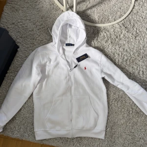 Vit hoodie från Polo Ralph Lauren - Vit hoodie från Polo Ralph Lauren med dragkedja och huva. Klassisk design med röd broderad logga på bröstet och två fickor framtill. Tillverkad i mjuk bomull, perfekt för en clean och stilren look.