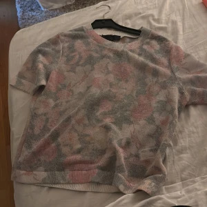 Blommig stickad tröja i pastell - Superfin stickad tröja med kort ärm och blommigt mönster i mjuka pastellfärger som rosa, grått och beige. Rund halsringning och ribbade kanter ger en chill vibe. Perfekt för dig som gillar romantiska och avslappnade looks.