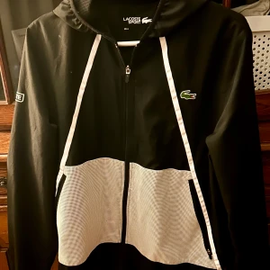 Svart vindjacka från Lacoste Sport Storlek M - Svart vindjacka från Lacoste Sport med vit meshpanel nedtill och vita detaljer längs dragkedjan. Jackan har huva, lång ärm och klassisk Lacoste-logga på bröstet samt patch på ärmen. Perfekt för dig som gillar sportig och clean stil.