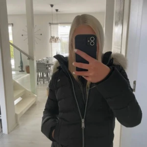 Svart dunjacka Tommy Hilfiger pälsluva - Svart dunjacka från Tommy Hilfiger med fluffig fuskpäls på luvan. Jackan har quiltad design, dragkedja framtill och två fickor. Klassisk logga på bröstet och snygg passform som håller dig varm under kalla dagar.
