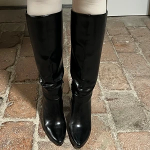 Svarta höga stövlar med klack - Säljer ett par svarta höga stövlar med snygg spetsig tå och bred klack. Stövlarna är tillverkade i blankt skinn och har dragkedja på insidan för enkel påtagning.