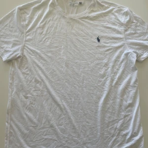 Vit t-shirt från Polo Ralph Lauren - Klassisk vit t-shirt från Polo Ralph Lauren med blå broderad logga på bröstet. T-shirten har rund halsringning och är tillverkad i mjuk bomull. Perfekt basic-plagg med normal passform och korta ärmar. Har en fläck som är svårt att se på bild 3