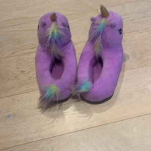 Lila enhörningstofflor med horn - Supergulliga lila tofflor formade som enhörningar med mjuk plysch, regnbågsfärgad man och svans samt glittrigt guldhorn. Perfekta för att mysa runt hemma och ge en färgklick till vardagen. Säljs i par.