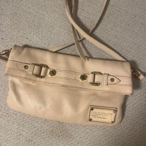 Beige axelväska Marc by Marc Jacobs - Snygg beige axelväska från Marc by Marc Jacobs i mjukt skinn med guldfärgade metalldetaljer och dekorativa spännen. Väskan har några fläckar som man ser på bilden, ingen man tänker på. Går möjligtvis att tvätta bort!😊nypris runt 2000kr.