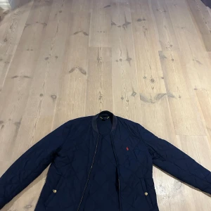 Mörkblå quiltad höst/vår jacka Polotröja Ralph Lauren - Snygg mörkblå quiltad höst/vår jacka från Polo Ralph Lauren med gulddetaljer på fickorna och klassisk röd logga på bröstet. Jackan har lång ärm, ribbad krage och dragkedja framtill. Perfekt för dig som vill ha en stilren och sportig look. Det står i taggen att den är storleken xl men skulle säga mer som xs. Passar personer som är 155-175 skulle jag säga