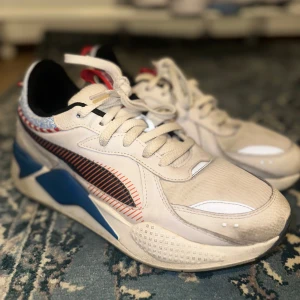 Puma RS-X 'Overnight Delivery' sneakers - Säljer ett par feta Puma RS-X sneakers i vitt, blått och rött med coola detaljer som 'Overnight Delivery' på hälen. Skorna har snörning, chunky sula och mönstrade partier i blått och rött. Perfekta för dig som vill sticka ut med en unik streetstyle.