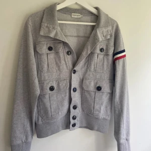 Grå kofta från Moncler med fickor - Snygg grå kofta från Moncler med fyra stora fickor framtill och knappar hela vägen. På ena ärmen finns Moncler-logga och ränder i blått, vitt och rött. Mjuk bomullskänsla och klassisk krage. Perfekt för dig som gillar street och sportig stil.