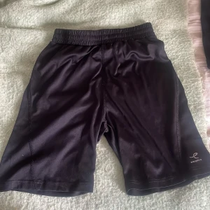 Svarta träningsshorts från Energetics - Svarta träningsshorts från Energetics med elastisk midja och snörning för perfekt passform. Materialet är mjukt och lätt polyester som andas bra. Shortsen har en enkel design utan mönster och loggan syns diskret på benet. Perfekta för gymmet eller löprundan.