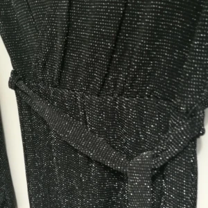 Svart glittrig långklänning med bälte - Svart långklänning med glittriga detaljer och smala vita prickar. Klänningen har lång ärm, v-ringning och ett matchande bälte i midjan. Materialet är stretchigt och ger en snygg passform. Perfekt för dig som vill sticka ut med en elegant look.