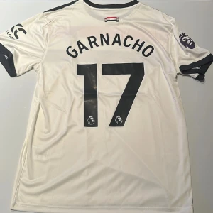 Manchester United Garnacho tröja Adidas - Manchester Uniteds officiella bortatröja med Garnacho 17 på ryggen. Notera bild 2, trycket ser ut att ha lite bubblor på sig, men det påverkar inte trycket. Därav priset. Vit bas med svarta och röda detaljer, svarta ränder på axlarna och svarta muddar. Adidas-logga, Snapdragon sponsortryck och Premier League-märke på ärmen. Tillverkad i lätt och ventilerande polyester. Replika. Endast handtvätt gäller!