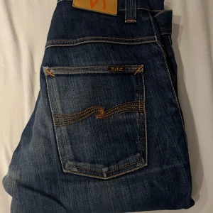 Mörkblå jeans från Nudie Jeans - Säljer ett par mörkblå jeans från Nudie Jeans med klassiska orangea sömmar och snyggt broderad bakficka. Jeansen har en rak passform och är tillverkade i slitstarkt denimtyg av bomull. Perfekta för dig som gillar stilrena och tidlösa jeans. W28 L32, tror det är lean Dean för att lappen är bort tvättad 