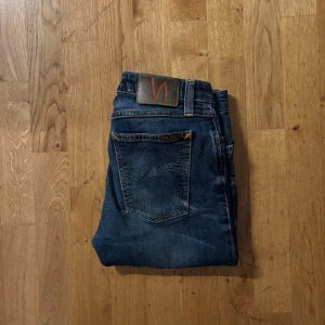 Nudie Jeans skinny blå jeans - Snygga blå jeans från Nudie Jeans med skinny passform och klassiska orange sömmar. Jeansen har slitna detaljer och råa kanter vid bensluten för en cool look. Tillverkade i mjukt denim med broderad bakficka och läderpatch med logga.
