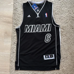 Svart Miami Heat baskettröja Adidas S - Snygg svart baskettröja från Adidas med Miami-tryck och nummer 6 på framsidan. Tröjan är ärmlös och har vita detaljer längs kanterna samt broderad NBA-logga. Tillverkad i ventilerande polyester, perfekt för sport eller streetwear. Köpt för 1099, lappen är kvar och är ej använd