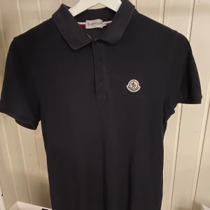 Svart pikétröja från Moncler - Svart pikétröja från Moncler med klassisk krage och knäppning framtill. Tröjan har korta ärmar och Monclers ikoniska logga broderad på bröstet. Tillverkad i mjuk bomull som känns skön mot huden. Osäker på hur äkta den är. Perfekt för en clean och stilren look.