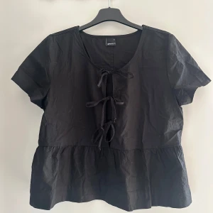 Svart blus med snörning  - Svart blus från Gina Tricot med korta ärmar och volang nedtill. Toppen har snygg snörning framtill med tre knyt och en rundad halsringning. Aldrig använd. 