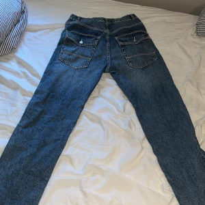 Blå raka jeans från Lindex, stl 164 - Säljer ett par klassiska blå jeans från Lindex i storlek 164. Jeansen har raka ben, stora bakfickor med knappdetalj och normal passform. Tillverkade i bomull med snygg tvättad look, perfekta för en avslappnad stil.