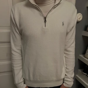 Vit half zip-tröja från Polo Ralph Lauren - Vit långärmad tröja från Polo Ralph Lauren med half zip-dragkedja och klassisk broderad logga på bröstet. Tröjan är i bomull och har en stilren, normal passform med ribbade muddar vid ärmslut och nederkant. Perfekt för en clean och snygg look. 
