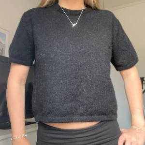 Svart stickad tröja med kort ärm - En svart stickad tröja med korta ärmar och rund hals. Tröjan har en enkel och stilren design, perfekt för lager-på-lager eller att bära som den är. Mjuk och skön känsla, passar till många olika outfits. Storlek 170