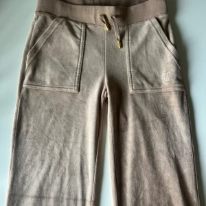 Beige mjukisbyxor Juicy Couture XS - Beiga mjukisbyxor från Juicy Couture med bred resår i midjan och snörning med gulddetaljer. Byxorna har två stora fickor fram och broderad JC-logga på ena fickan. Mjukt material som känns riktigt skönt mot huden.