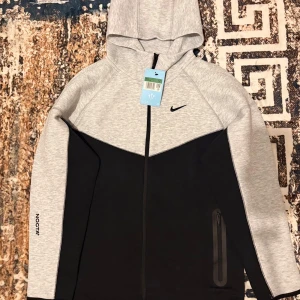 Nike NOCTA hoodie svart/grå - Nike NOCTA hoodie i svart och ljusgrått med hel dragkedja, huva och NOCTA-logga på ärmen. Snygg tvåfärgad design, långärmad och med praktisk ficka med dragkedja. Perfekt för en sportig och clean look. Storlek xl känns som L