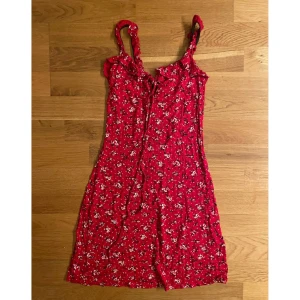 Röd blommig sommarklänning - Söt röd klänning med vita och svarta blommor, smala axelband och volangdetaljer upptill. Perfekt för varma dagar med sin korta längd och lätta material. Klänningen har en feminin och avslappnad stil som passar till många tillfällen.