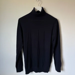 Mörkblå polotröja i merinoull från H&M - Klassisk mörkblå polotröja från H&M i mjuk merinoullsblandning. Tröjan har lång ärm, hög polokrage och en smal, stilren passform. Perfekt för lager-på-lager och enkel att matcha med allt. Snygg och tidlös basic för garderoben. 3V5