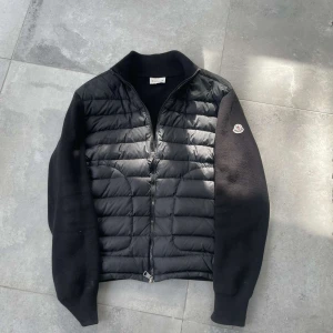 Svart pufferjacka från Moncler - Snygg svart pufferjacka från Moncler med quiltad front och stickade ärmar. Jackan har dragkedja framtill och Moncler-logga på ena ärmen. Perfekt för dig som vill ha en stilren och sportig look.
