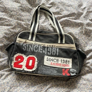 Svart axelväska med tryck och brodyr - Cool svart axelväska med vita detaljer, tryck och broderad röd siffra 20 framtill. Väskan har vita och svarta randiga handtag, dragkedja och flera fack. Perfekt för dig som gillar sportig och unik stil.