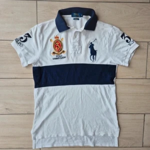 Vit och marinblå Polo Ralph Lauren t-shirt - Vit och marinblå t-shirt från Polo Ralph Lauren med broderad logga och emblem på bröstet, samt stora siffror på ärmarna. Klassisk pikékrage och bred marinblå rand över magen. Tillverkad i bomull och har en normal passform.